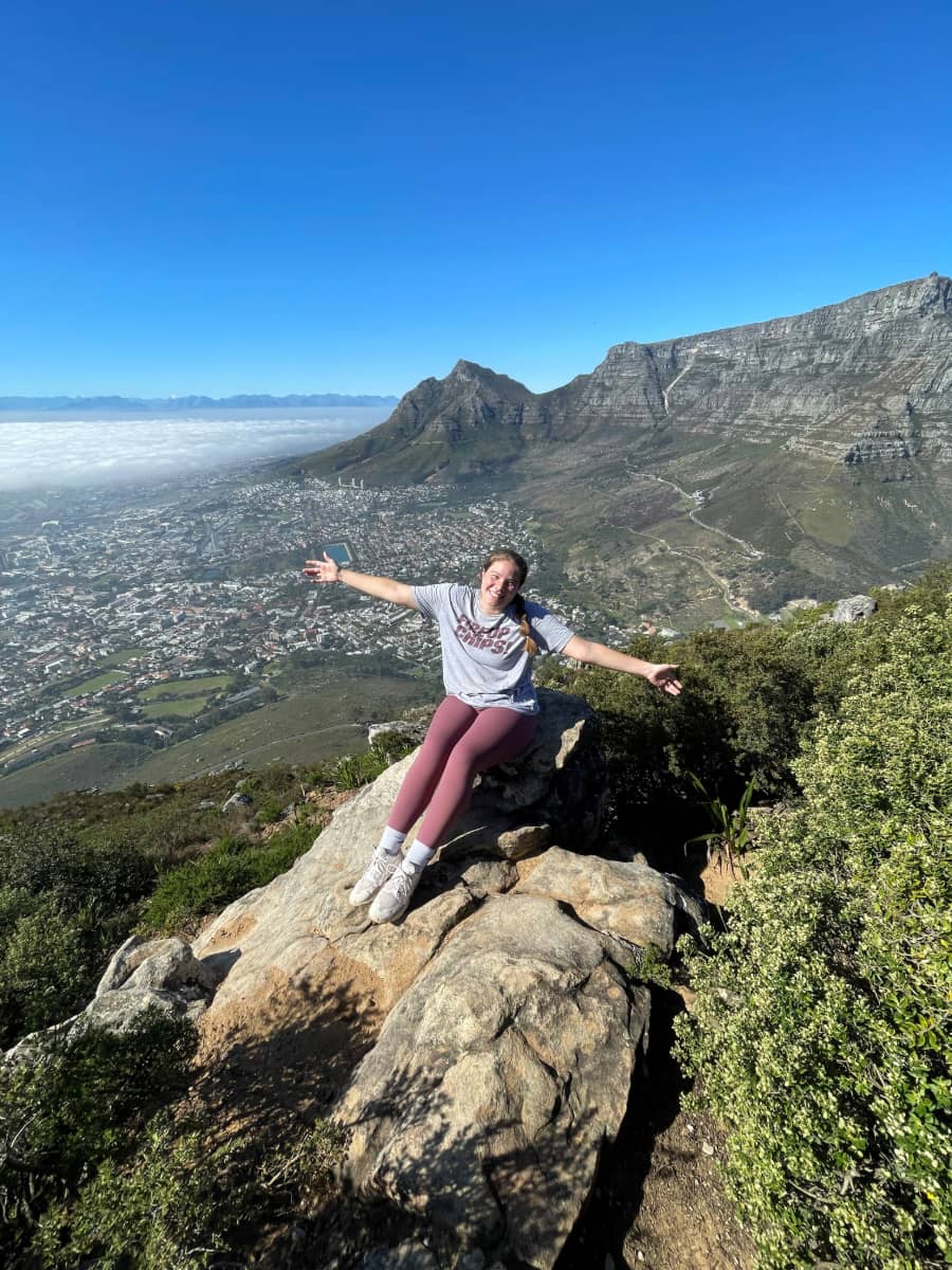 A study abroad student sitting on a rock with their arms outstretched, overlooking a vast landscape of mountains and a city below under the clear blue sky.