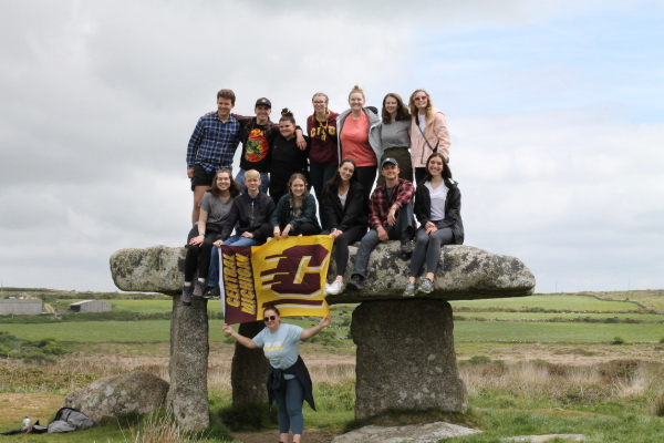 Study Abroad students sitting on a large, ancient stone structure in an open field holding a Central Michigan flag.