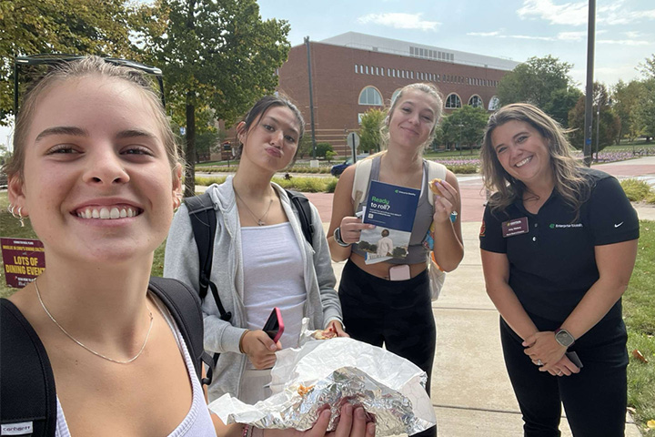 Abby Suchy stands with three other women on campus. She's holding foil with food in her hand and taking the photo as a selfie. The others are smiling and have phones and food.