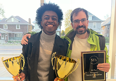 Professor and student pose with the trophies they earned at a debate tournament.