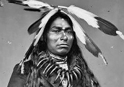Black and white image portraying a Chippewa warrior with face paint, a bear claw necklace and feathers adorning his head.