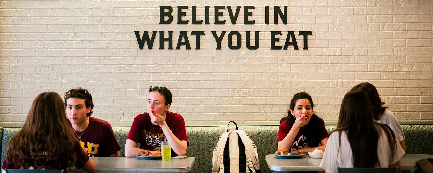 Six students sit in booths at the Eatery in Towers Hall, Central Michigan University, eating and chatting. A white brick wall behind them features the phrase