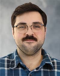 Jacob Helm, Interim Director of Sexual Aggression Services, professional headshot, dark hair, wearing a blue plaid button-down shirt, and glasses with a grey background.
