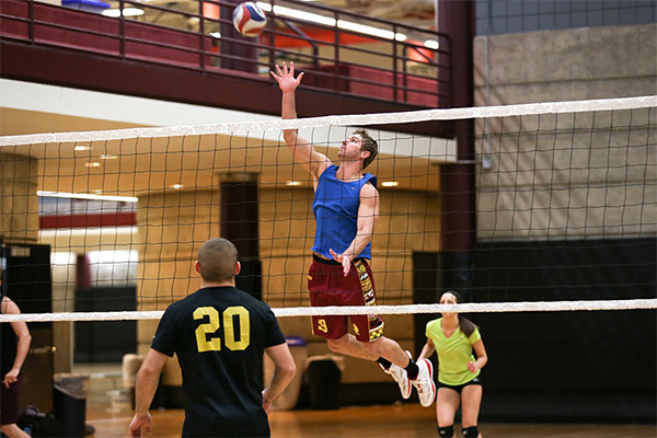 Male student wearing blue tank top and maroon shorts spiking a volleyball over the net