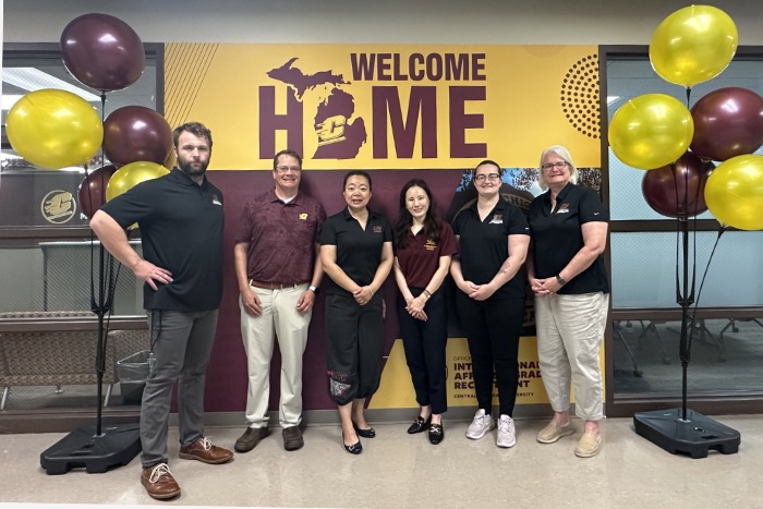 Office of International Students and Scholars group photo Six people stand in a line in front of a maroon and gold backdrop that says Welcome Home, with the state of Michigans outline replacing the O, and smile for a group picture in an office building. Maroon and gold balloons decorate the sides