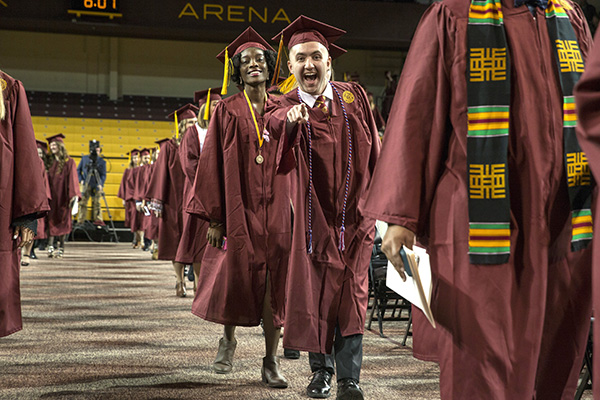 A student walking in the graduation ceremony pointing at the camera and smiling widely.