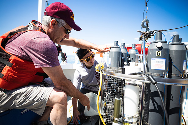 A CMUBS faculty member in a maroon hat and life jacket next to a student preparing deep water sampling equipment aboard a boat.