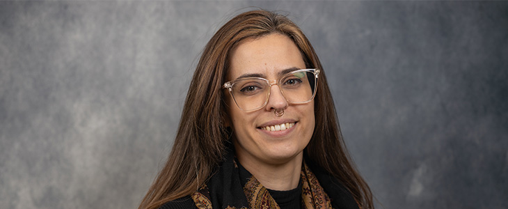 Professional headshot of Mariagabriella Stuardi in black top with earth tone scarf against a gray background.