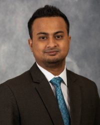 Image of Md Amin, a man, looking at the camera wearing a dark suit with white collar shirt, and blue tie, with grey background.