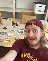 Image of Matthew Black, a man, wearing maroon shirt and cap, looking at the camera with photos his work desk for digitization.