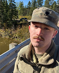 Still photo of graduating libraries student employee Larry Laponsie in beige hunting jacket and army green bucket hat against pine trees and tall grass wooded area