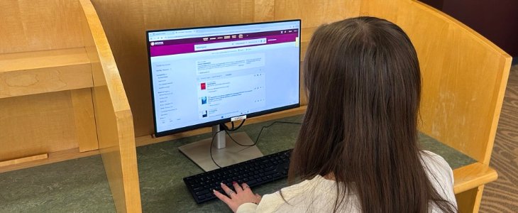 Image depicts a student sitting at a library computer, logged in searching the Library's Smart Search software. They are searching Hemmingway