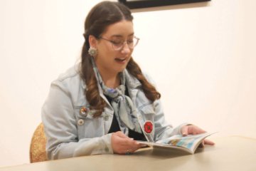 A young woman with brown hair sits at a table, reading a magazine. She wears glasses, a denim jacket with pins, a scarf, and earrings. She appears to be speaking or reading aloud with a slight smile.