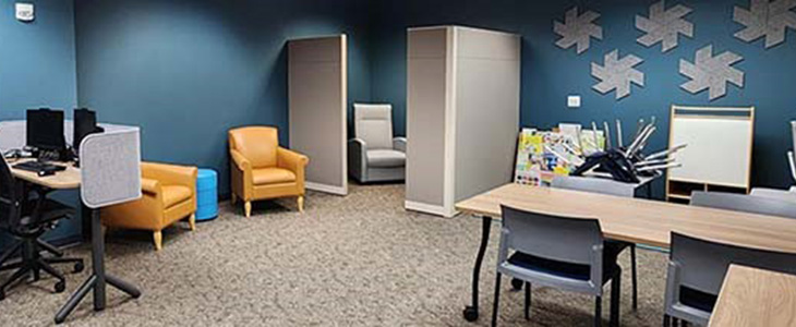 A room in the library with wall decorations, multiple desks and plush chairs for families to use.