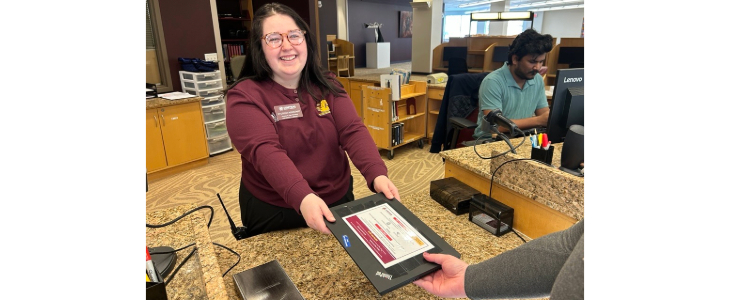 At the Park Library, Library Services Desk, an employee is seen handing a closed laptop to a student patron after the student checked it out as a loan.