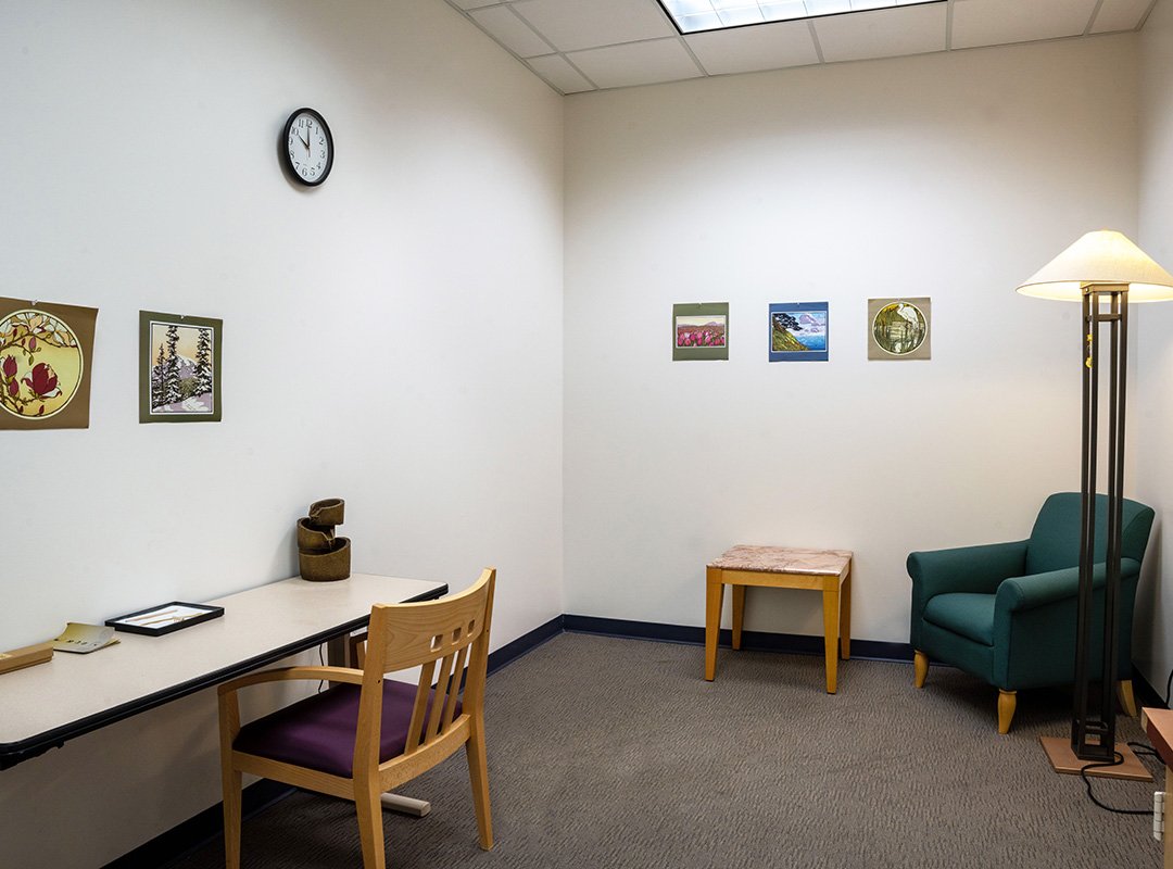 Image of two walls in the Quality of Life Room at the CMU Park Library.  Walls are white with a few pictures, furniture featured, slim table, wood chair with arm, end table, lamp, and cushioned green chair