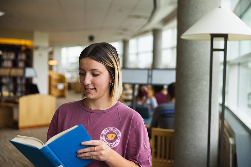 student holding book student wearing maroon and gold holds open a book and smiles while standing in the CMU Park Library