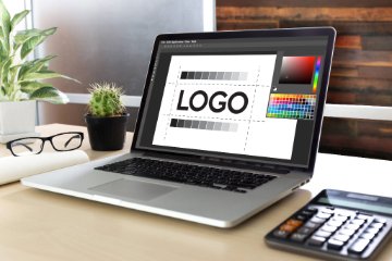 A laptop displays a logo design in progress, accompanied by a cactus, glasses, a rolled-up paper, and a calculator on a clean, organized desk.