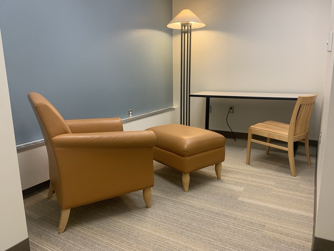 The image shows a small room with a brown leather armchair, matching ottoman, and a desk with a wooden chair. A floor lamp provides soft lighting. The walls are plain, one blue, and the space feels simple and calm.