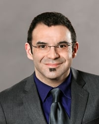 Joseph Michael Sommers wearing a gray jacket and tie with a blue shirt and smiling while posing for a professional headshot.