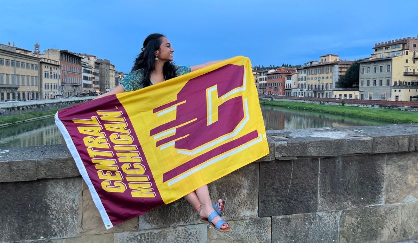 Student sits on a bridge in Florence, Italy holding a Central Michigan University flag and looking to the side.