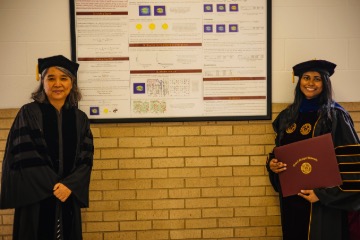 A student and faculty member standing in front of their research, both wearing graduation gowns.