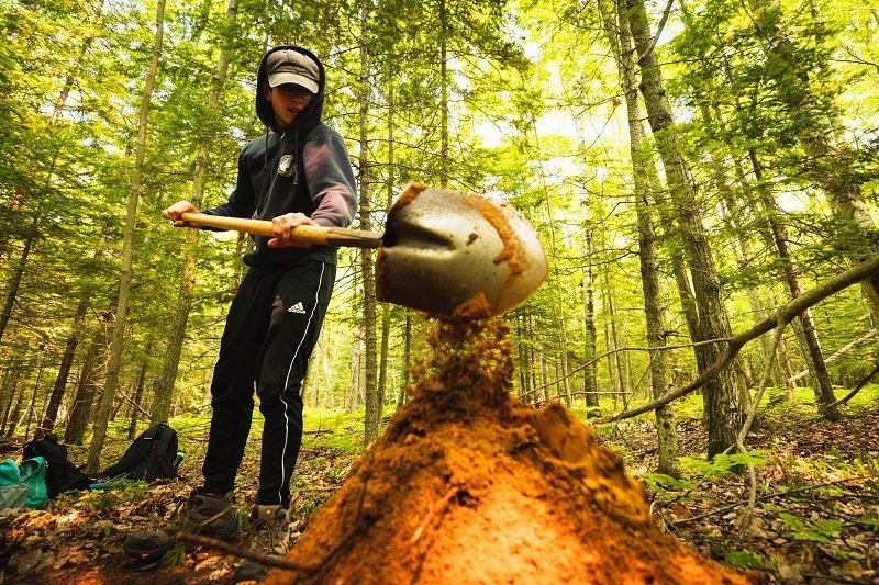 A student in the forest digging and putting the dirt in a pile.