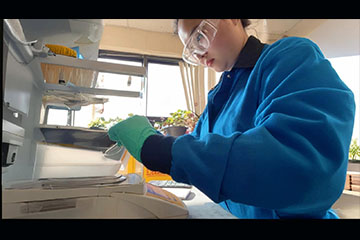 A woman wearing safety goggles, gloves and a lab coat works in a research lab on campus.