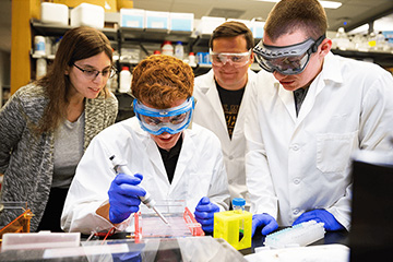 A group of people in lab coats and goggles with a woman looking over their shoulders.