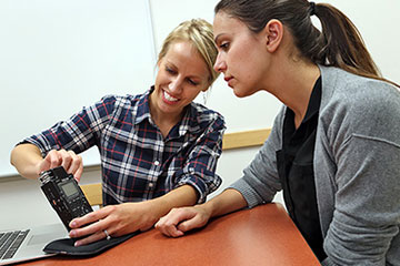 Two women sitting near one another. A woman with blonde hair shows a student with long dark hair a recording device.