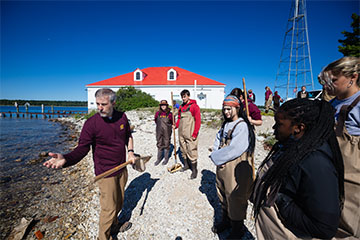 A group of students in waders stand on a rocky beach with a CMU faculty member.