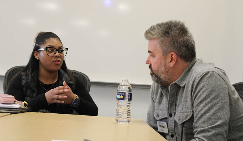 A student speaking to an NVC mentor at a table in a classroom.