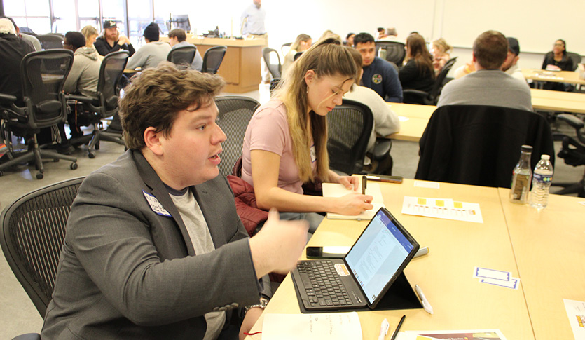 Two students speaking and writing at a table in a classroom.