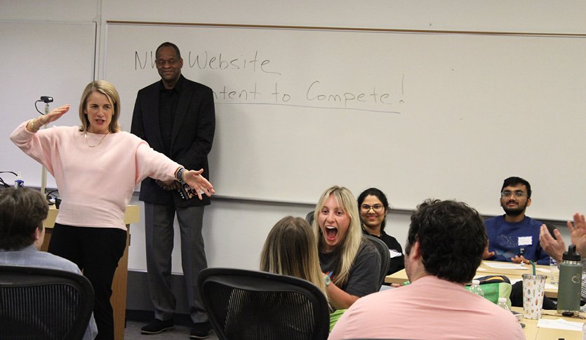 A professor pitching an idea in front of a group of smiling students.