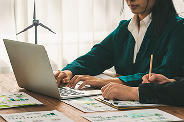 A woman in a teal blazer types on a laptop surrounded by environmental reports and a model of a wind turbine.