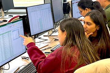 Two female students sit in front of computer monitors while one in a red sweatshirt points to data that appears on the screen.