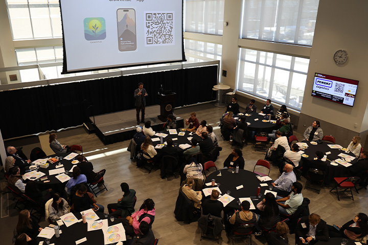 Students sit at round tables in the Grawn Atrium during the 2025 Ethics Summit. They are watching a speaker under a drop down screen. They have worksheets in front of them.