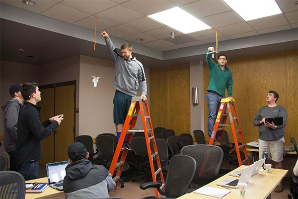 Lean Six Sigma workshop with students testing the flight of paper helicopters. Two students stand on ladders as they drop the paper to the ground. Four other students observe with stop watches and record the time.