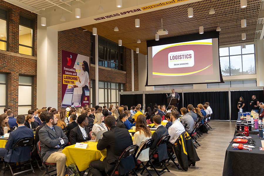 A speaker at the front of a large, open room addresses event attendees in business attire sitting at long tables