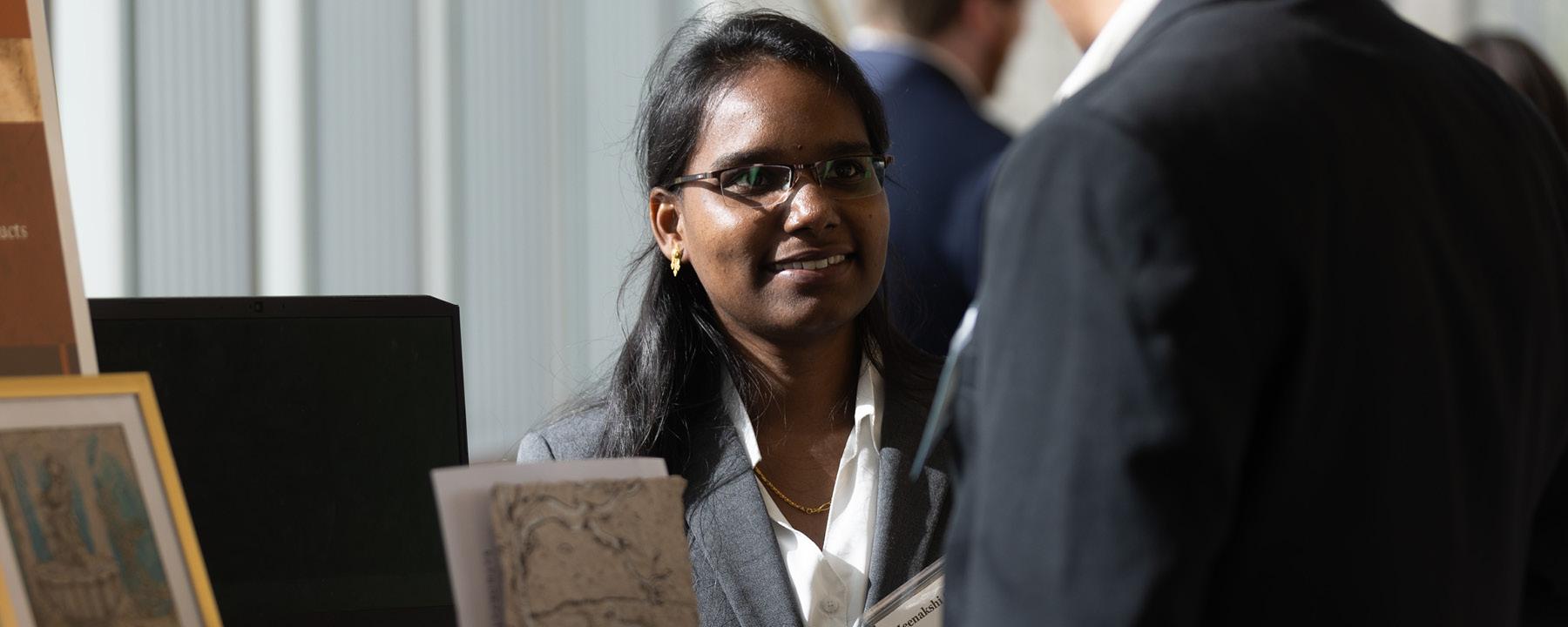 A student in a suit standing next to a table smiling at another person.