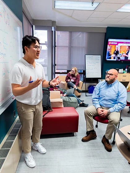 A man in a white polo hold a dry erase marker while standing in front of a white board talking to a classroom audience.