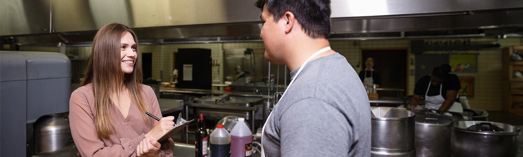 Female hospitality student holding a clipboard while talking with a male chef in a hotel kitchen.