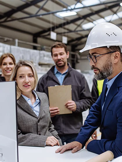 A group of female and male logisticians look at a computer screen while standing in a warehouse.