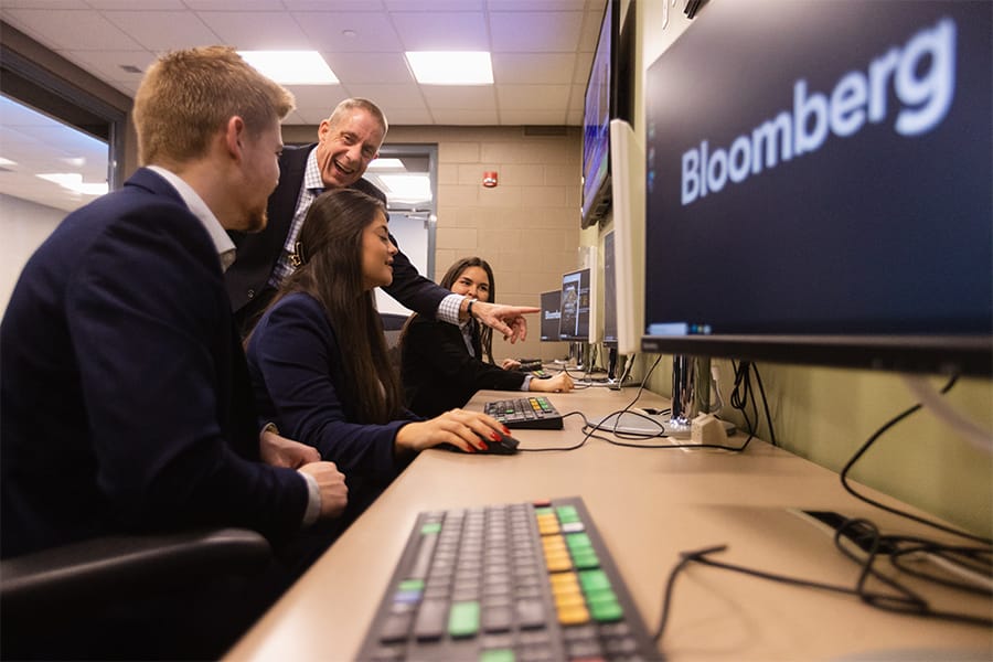 A group of finance students dressed professionally works with a professor on the Bloomberg terminals.