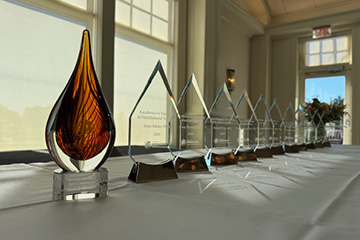 A row of glass awards sit on a long white table in front of a window.