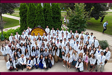 A group of smiling medical students gathered outdoors around a sign displaying the University seal.