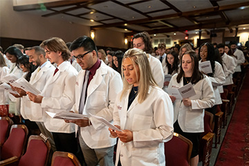 Group of young men and women in white coats read from booklets in an auditorium.