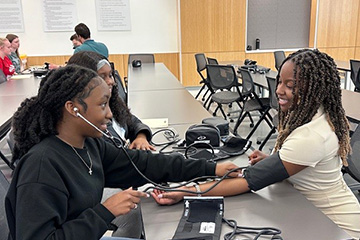 Three female students in a classroom, sitting at a table learning how to check blood pressure.