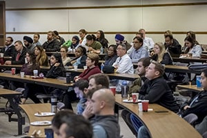 Medical students sit at tables in a lecture room.