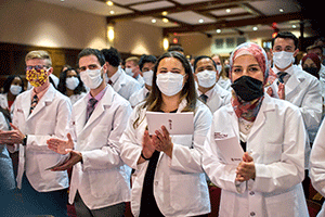 A group of medical students wearing white lab coats and face masks stand together and clap.